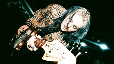 lleo lleo, with blond and gray hair, wearing a Burberry shirt and playing the guitar. She has dark eye make up and looks directly at the camera